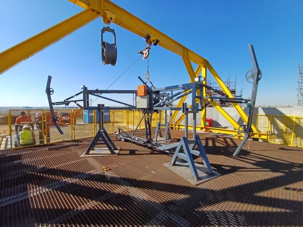 A metal testing rig on a grated platform under a bright blue sky. The setup includes a yellow gantry crane and multiple metallic components and wires. Construction materials and workers are visible in the background.