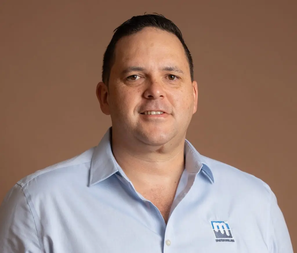 A man with short dark hair wearing a light blue collared shirt with a blue and white embroidered logo stands in front of a plain beige background, looking at the camera and smiling slightly.