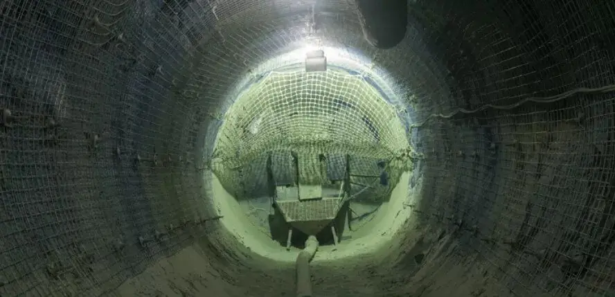 A dimly-lit underground tunnel reinforced with metal netting on the walls and ceiling. The floor is rough and appears to have construction equipment and debris scattered around, suggesting an ongoing or recently completed excavation project.