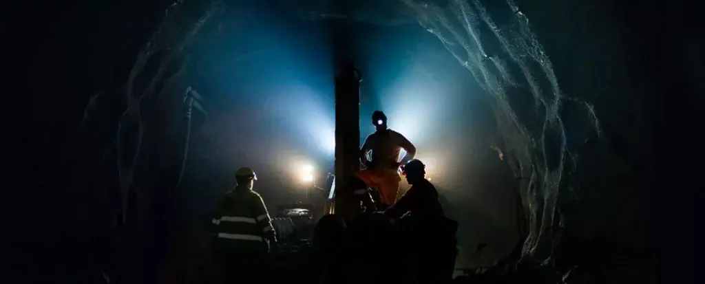 Three miners in safety gear work underground in a dimly lit tunnel. One miner stands and operates machinery illuminated by blue lights, while two others observe. The rocky tunnel walls encompass the scene, adding to the sense of depth and enclosed space.