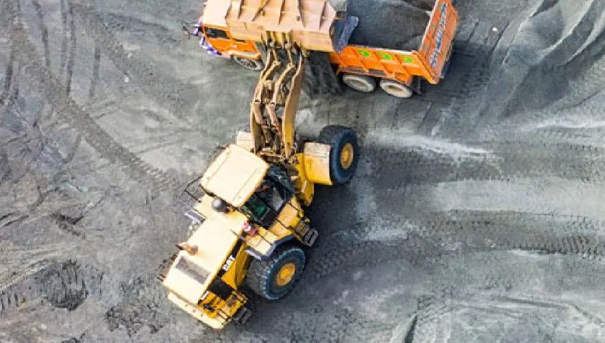 Uma vista aérea de um canteiro de obras mostra uma escavadeira amarela carregando terra em um caminhão basculante laranja. Os veículos estão em uma superfície irregular, cinza e de terra, com marcas de pneus visíveis ao redor deles. O braço da escavadeira está estendido na traseira do caminhão basculante.