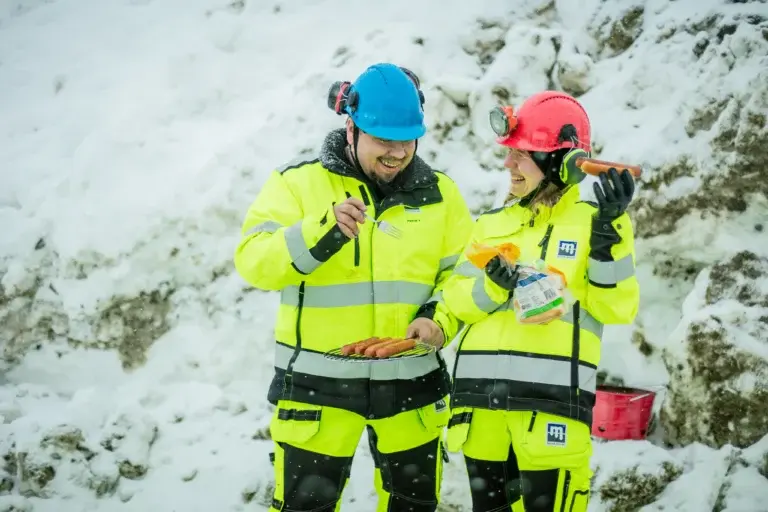 Dois trabalhadores com equipamentos de segurança, capacetes e luvas amarelos brilhantes estão fazendo uma pausa em uma paisagem com neve. Um deles segura um garfo e um prato com salsichas, enquanto o outro segura pão e salsichas. Eles parecem estar tendo uma conversa descontraída.