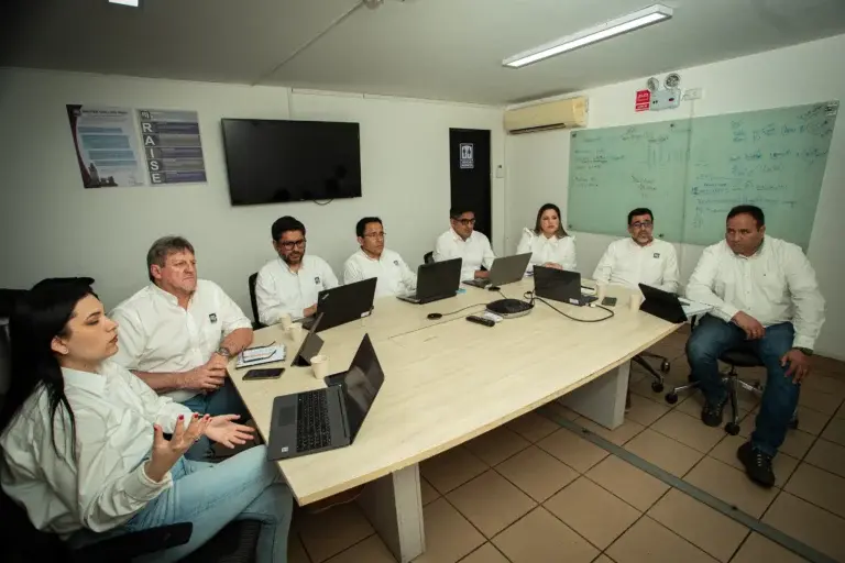 Um grupo de oito pessoas sentadas ao redor de uma mesa de conferência com laptops à sua frente, envolvidas em uma discussão. Elas estão em uma sala com um quadro branco cheio de anotações e diagramas, uma TV na parede e um pôster na parede. Todos estão vestidos com camisas brancas.