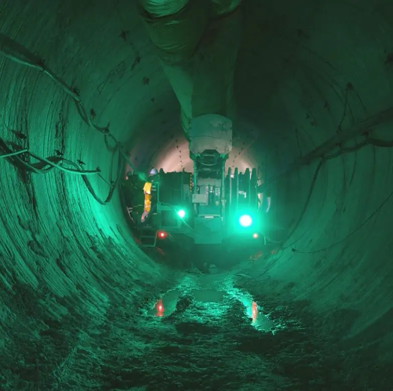 Um trabalhador opera um maquinário dentro de um túnel grande e iluminado com paredes irregulares e irregulares. A iluminação verde emite um brilho sinistro e há uma pequena poça de água no chão que reflete as luzes. A cena parece industrial e subterrânea.