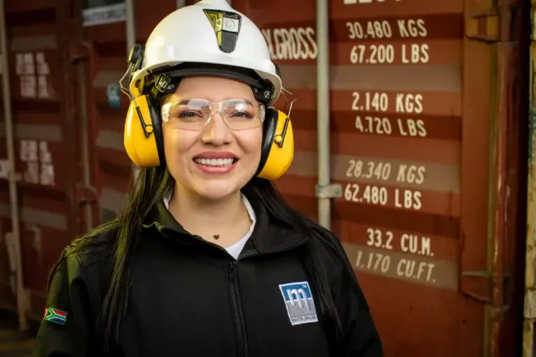 Uma mulher vestida com equipamentos de segurança, incluindo um capacete branco, protetores auriculares amarelos e óculos de segurança, está sorrindo em frente a um contêiner de transporte. Ela está vestindo uma jaqueta preta com um logotipo com as letras "mi". O contêiner tem várias marcações de peso.