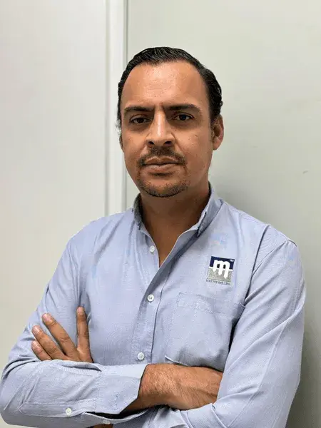 A man with short dark hair and a trimmed beard is standing against a plain wall with his arms crossed. He is wearing a light blue button-up shirt with a logo on the left chest that appears to be a stylized “m”. He has a neutral expression and is looking at the camera.
