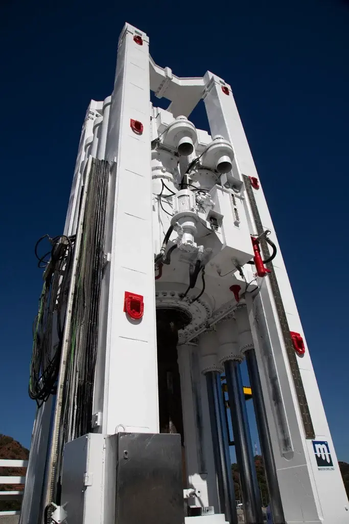 Uma máquina robótica vertical e branca com componentes mecânicos complexos, cabos e detalhes em vermelho se destaca contra um céu azul claro. A máquina apresenta vários cilindros, engrenagens e vigas estruturais, e está posicionada ao ar livre em um dia ensolarado.