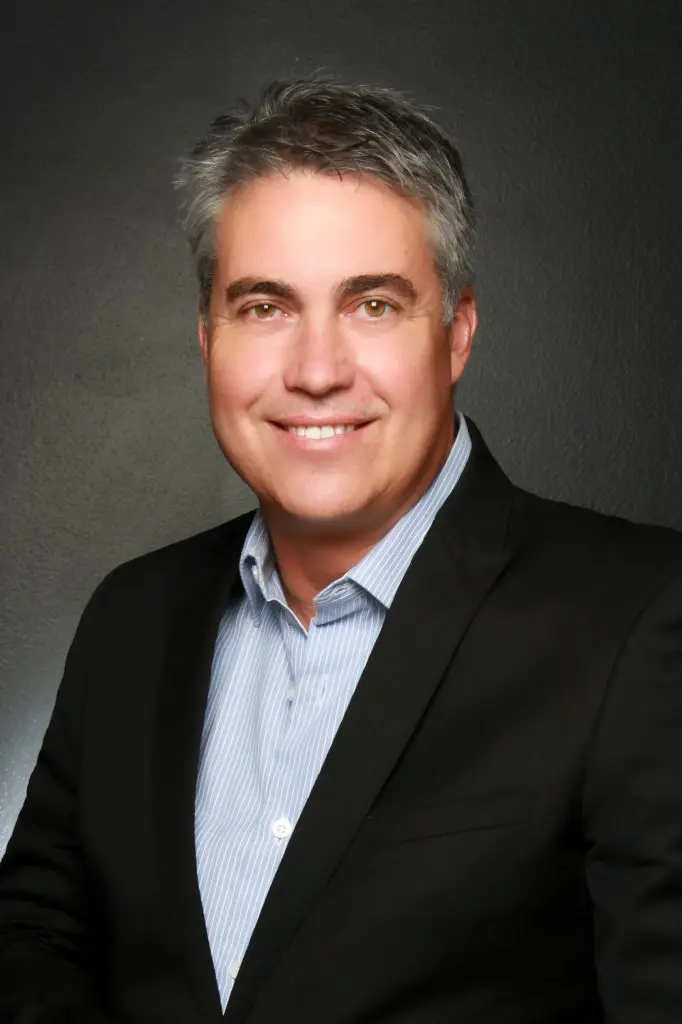 A man with gray hair wearing a black suit jacket and light blue striped shirt smiles at the camera against a dark gray background.