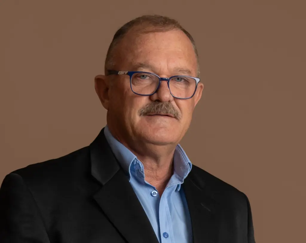 A middle-aged man with short hair, glasses, and a mustache is wearing a black suit jacket over a light blue shirt. He is posing against a plain brown background.
