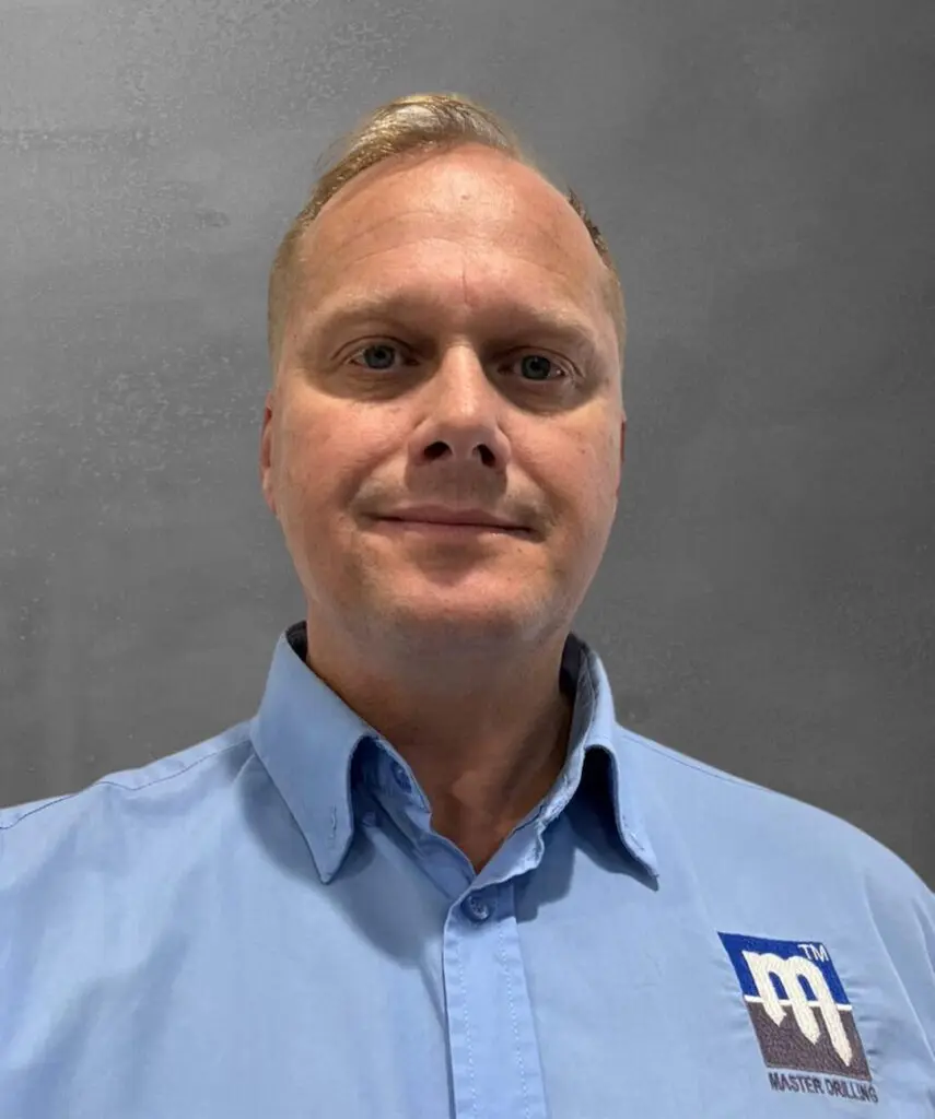 A man with short blonde hair is wearing a light blue collared shirt featuring the logo of “Master Drilling” on the left side. He is posed against a plain gray background and looking straight at the camera with a neutral expression.