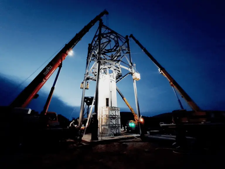 A group of cranes erects a large metal framework during dusk. The sky is dark blue, and the cranes' lights illuminate the structure and surrounding area. Workers and equipment are visible at the base of the framework.