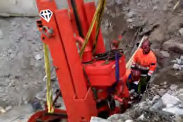 A worker in a safety vest operates a large, red construction drill amidst a rocky excavation site. Surrounding the worker and machine is a grey, rough terrain, suggesting ongoing construction or geological work.