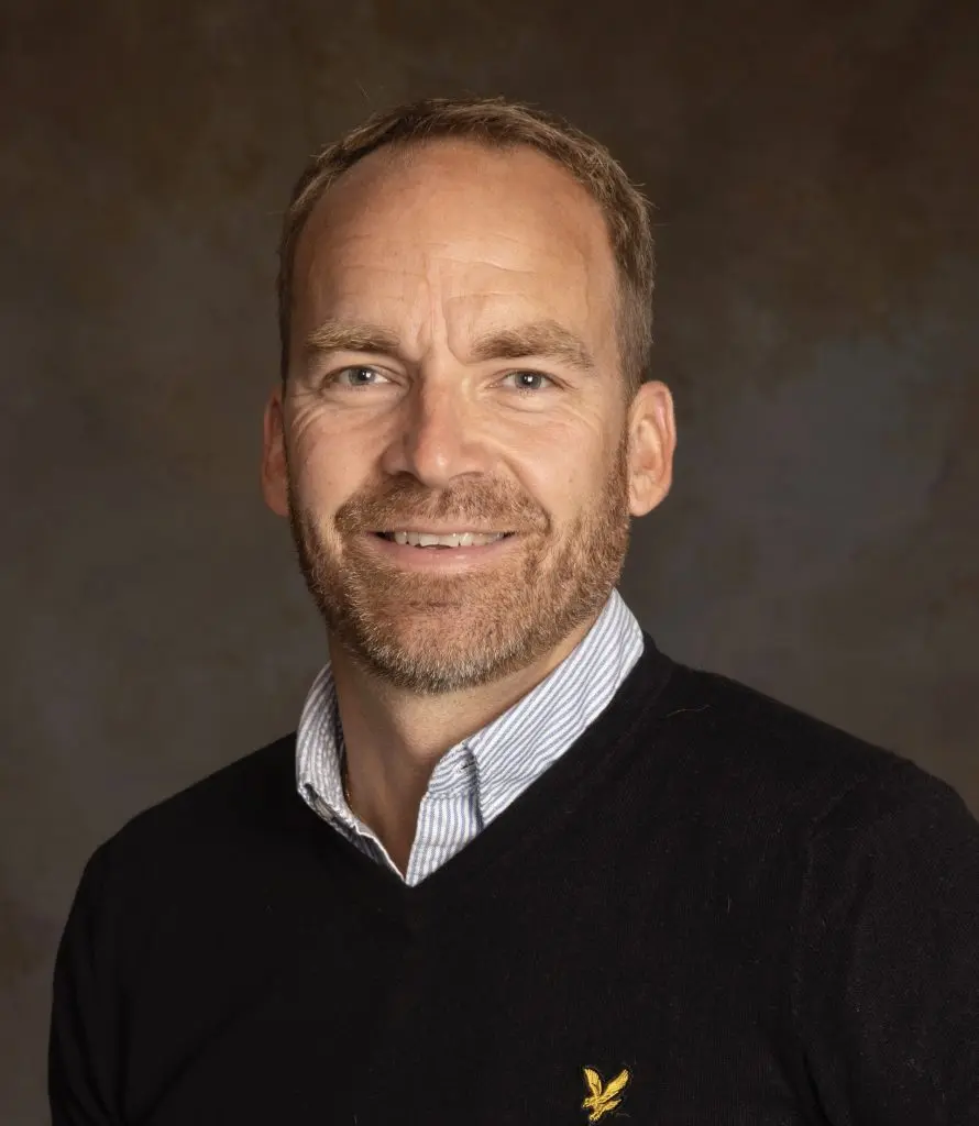 A man with light brown hair and a beard, wearing a black sweater over a striped collared shirt, smiles at the camera against a brown, textured background.