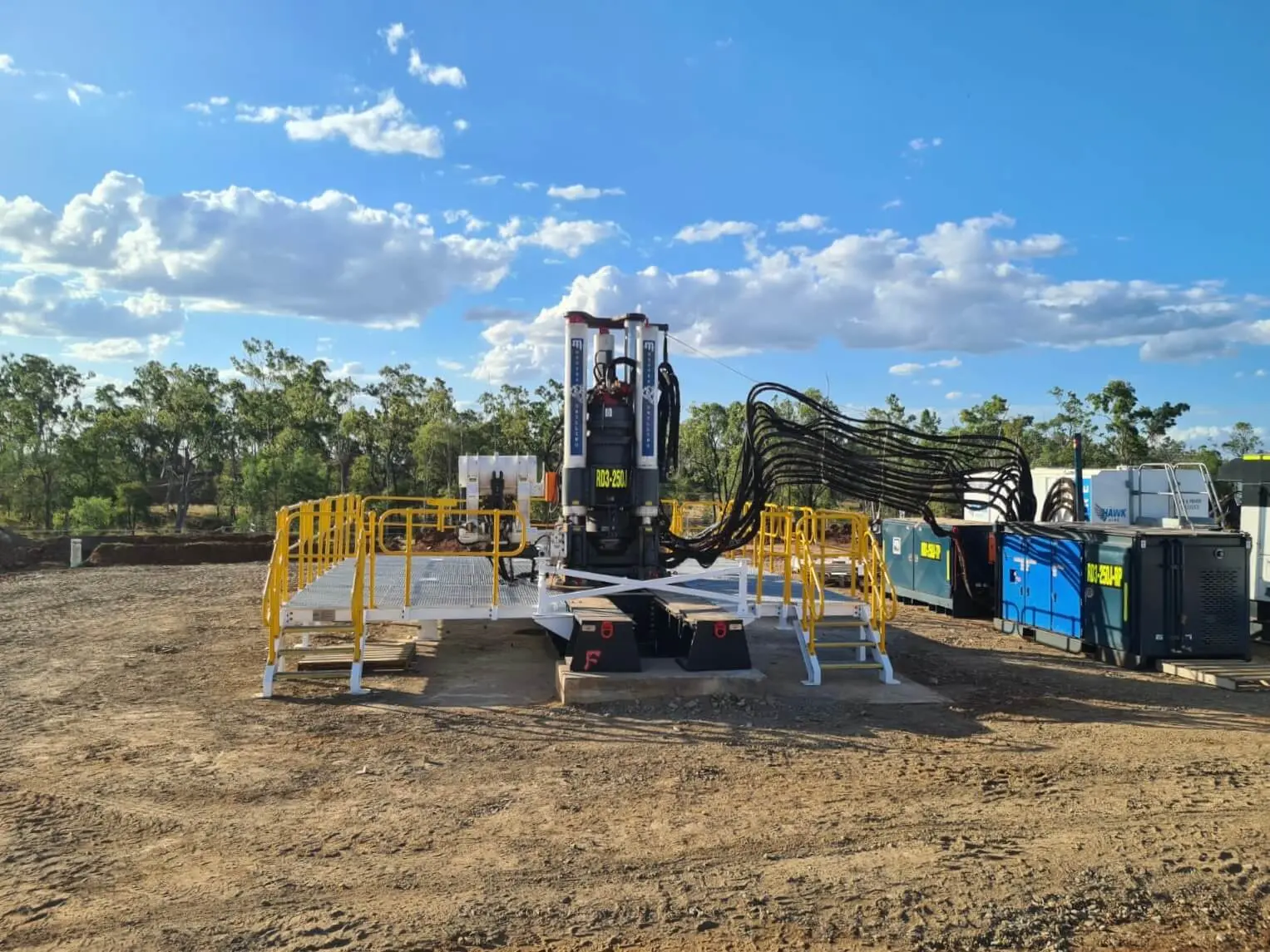 An RD3-250J raise drill rig stands on a white platform with yellow handrails on a cleared bushland site under a blue sky.