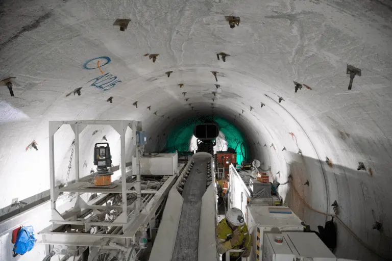 A wide tunnel under construction features various heavy machinery and equipment. Workers in safety gear are seen within the tunnel, focusing on their tasks. The tunnel wall is marked with some graffiti and is well-lit, giving a clear view of the construction process.
