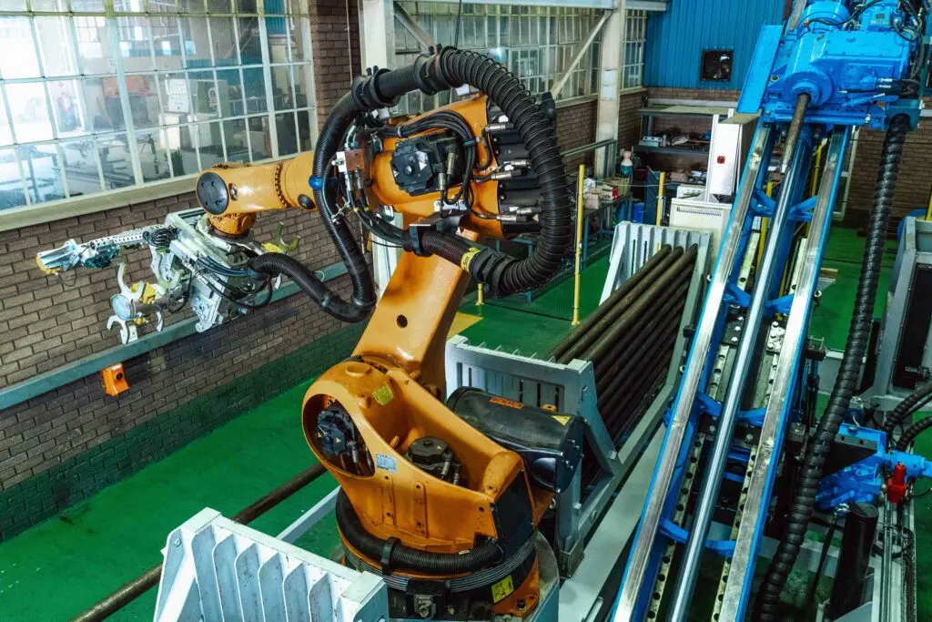 Um braço robótico em uma fábrica está manuseando longas hastes de metal. O braço está montado em uma plataforma ao lado de um sistema de transporte. O plano de fundo inclui grandes janelas e equipamentos industriais.