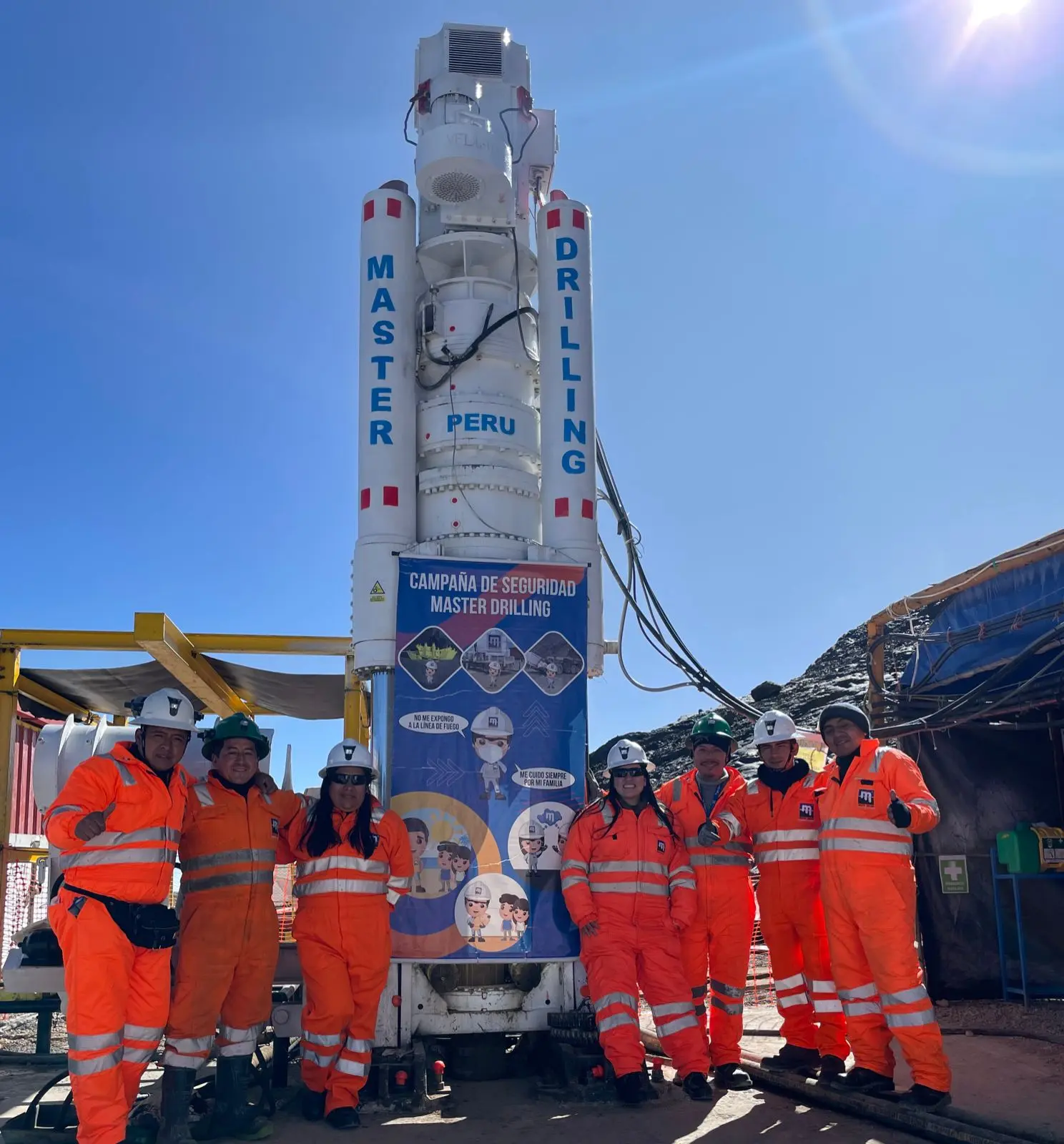 Sete membros da equipe com macacões laranja fazem sinal de positivo ao lado de uma perfuratriz branca da Master Drilling Peru com um banner de campanha de segurança.