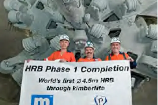 Three workers in orange safety uniforms and white hard hats are holding a banner that reads "HRB Phase 1 Completion. World's first Ø 4.5m HRB through kimberlite." They are standing in front of a large piece of industrial machinery with gears and components.