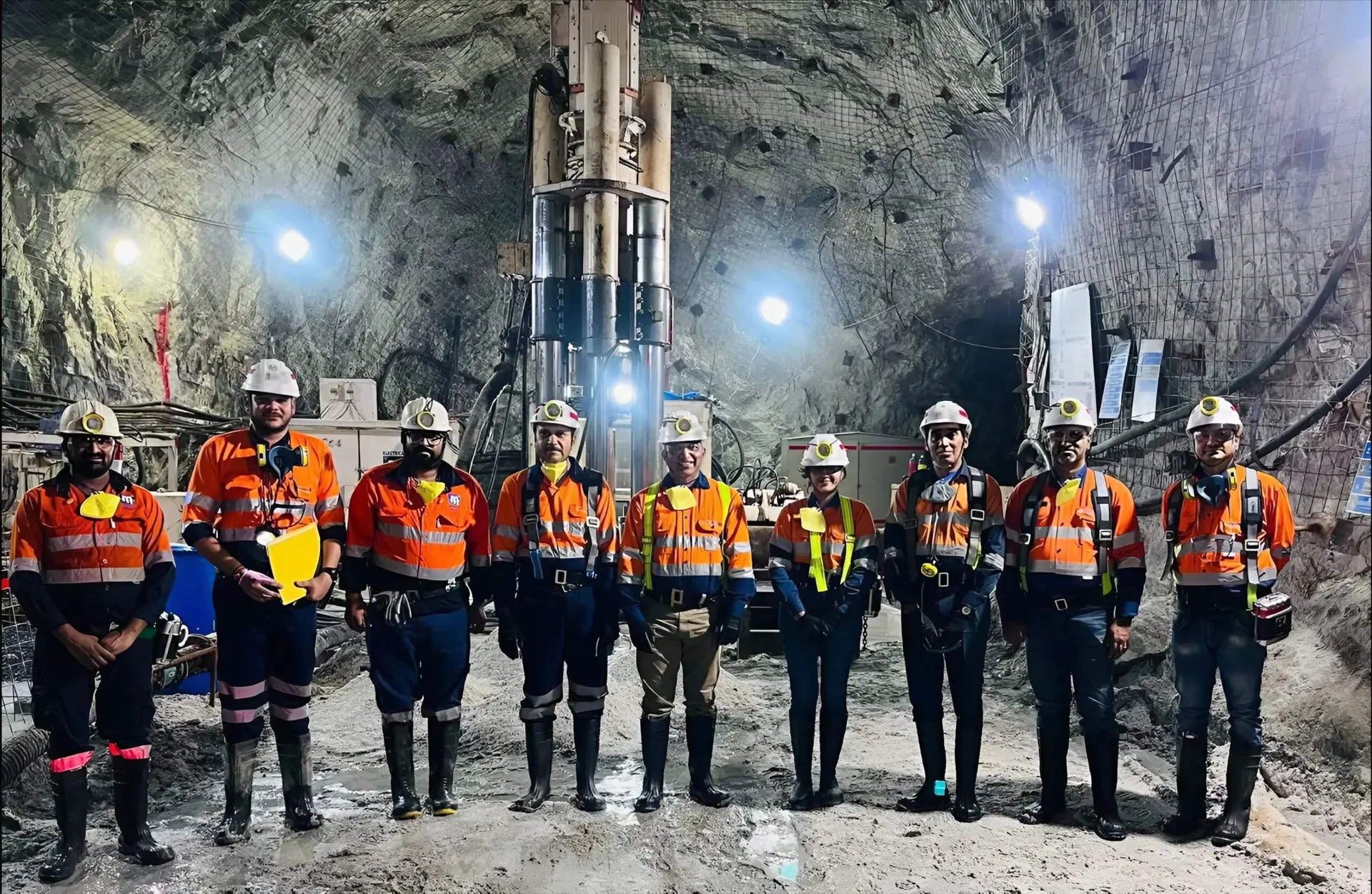 Nine crew members in orange safety wear line up in front of an underground raise bore rig in a mesh-bolted cavern.