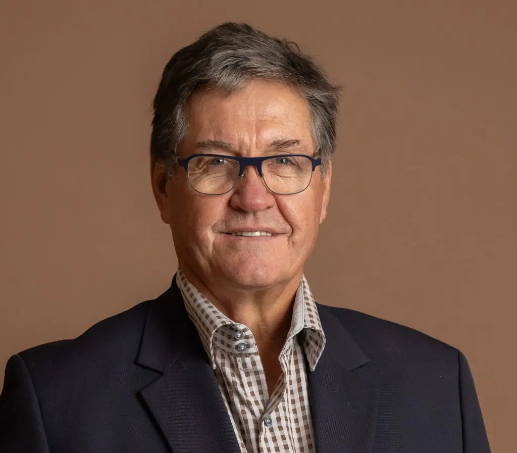 A middle-aged man with short gray hair, wearing glasses, a dark blazer, and a checked shirt, smiles slightly against a plain brown background.