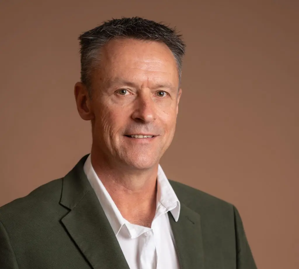 A man with short dark hair wearing a green blazer and white shirt smiles slightly against a plain brown background.