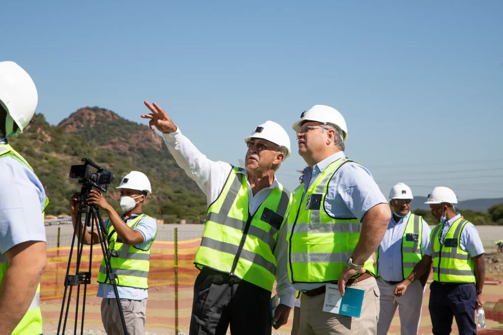 A group of construction workers wearing hard hats and reflective vests stand outdoors at a worksite. One man is pointing towards the distance while speaking to another man. A camera operator films the scene. Hills and clear blue skies are in the background.