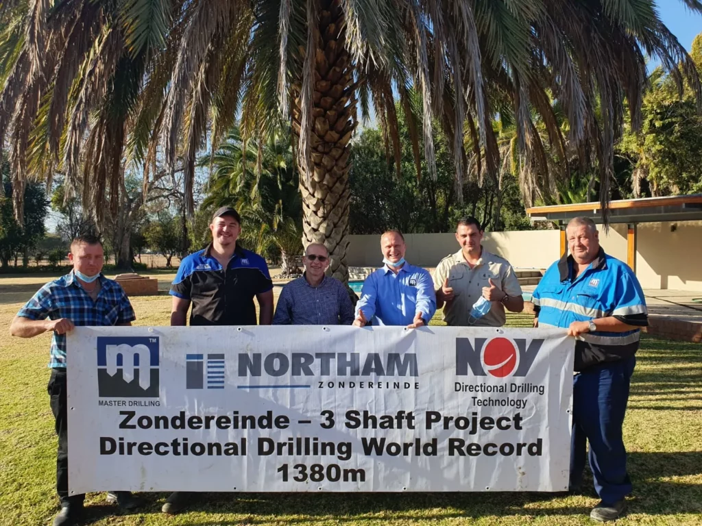 Six men stand behind a large banner reading, "Zondereinde - 3 Shaft Project Directional Drilling World Record 1380m." They are smiling, with palm trees and a building in the background.