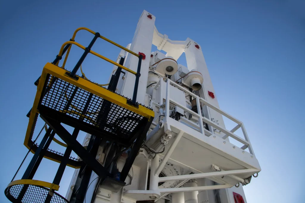 A low-angle view of a white industrial structure with stairs and railings against a clear blue sky. The complex metal framework includes platforms painted with black and yellow elements, and various pipes and components are visible.