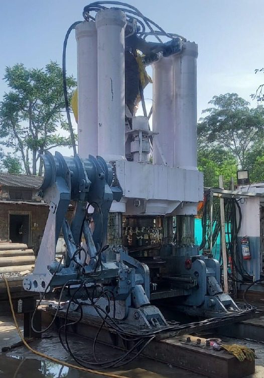 A large industrial machine with hydraulic components stands outdoors, surrounded by trees and construction materials. The metal structure features multiple pistons and hoses, with a blue and white color scheme. Ambient sunlight is visible.