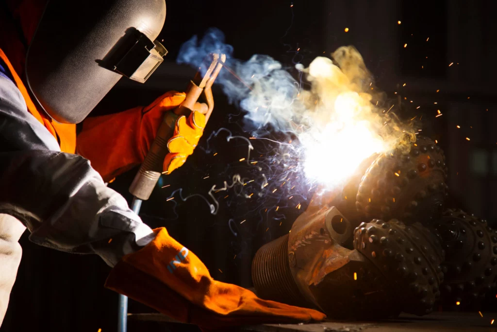 A person wearing a protective helmet and gloves is welding metal. Bright sparks and smoke are visible as the welding torch creates a glow. The area is dark, emphasizing the brightness of the welding process.