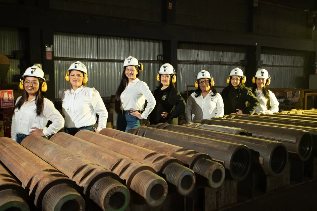 Six women wearing white helmets and safety earmuffs stand in a row inside an industrial facility. They are smiling and posing in front of large, cylindrical metal objects laid out on workbenches. The background shows industrial materials and equipment.