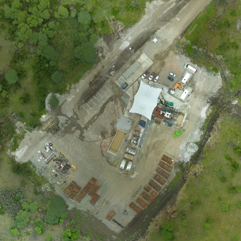 An aerial view of a large construction site nestled amidst greenery. The site seems organized with various sections including scattered vehicles, construction equipment, arranged stacks of materials, and an expansive white canopy structure at the center.