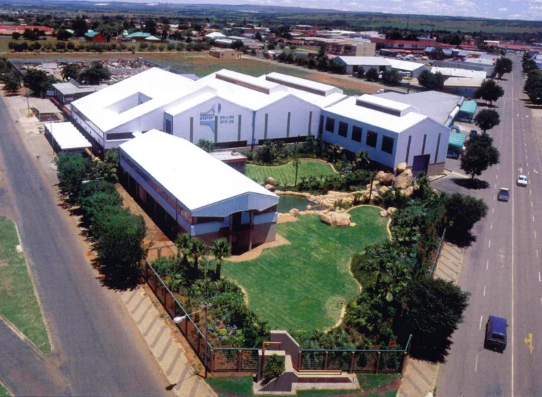 Aerial view of a landscaped, modern educational campus with multiple large white buildings, green lawns, gardens, and a playground. It is bordered by trees and situated next to a road with a few vehicles. The surrounding area shows more buildings and open fields.