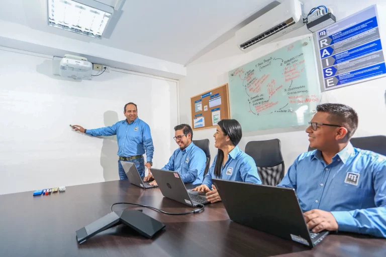 Four people in blue shirts sit at a table with laptops, while one of them stands and writes on a whiteboard. They appear to be engaged in a meeting or presentation in a modern office with notes and diagrams on the walls.