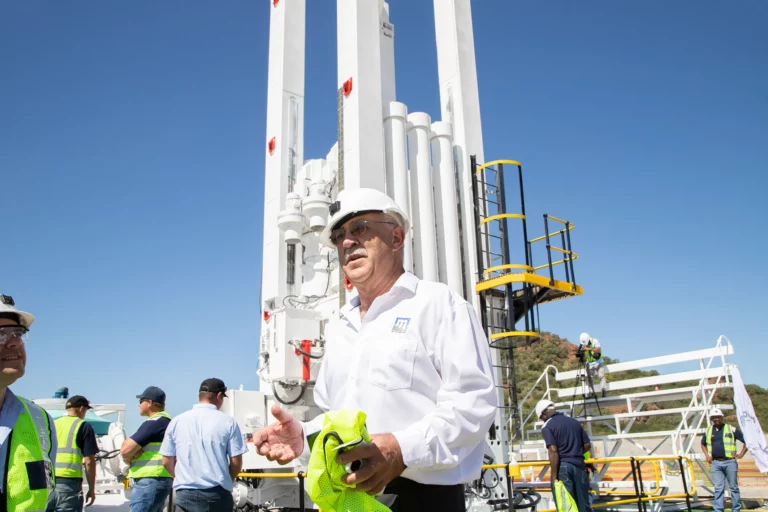 A man wearing a white safety helmet, sunglasses, and a white shirt with a logo stands in front of a large industrial structure outdoors. He holds a safety vest. Surrounding him, several people in yellow safety vests are engaged in various activities. The sky is clear and blue.