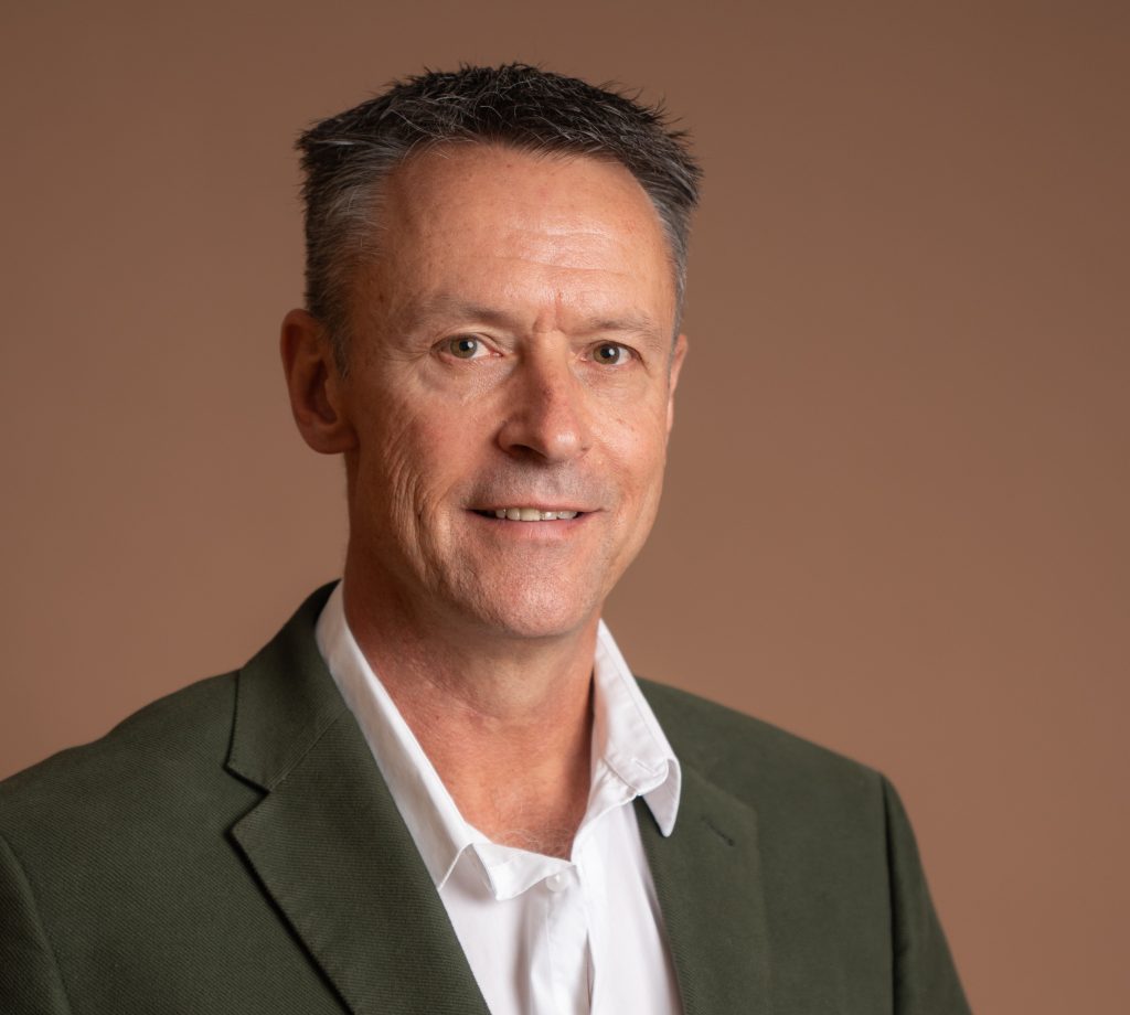 A man with short dark hair wearing a green blazer and white shirt smiles slightly against a plain brown background.