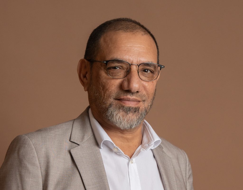A middle-aged man with glasses, a gray beard, and short hair wears a light gray suit jacket over a white shirt. He is smiling slightly and posed in front of a plain brown background.
