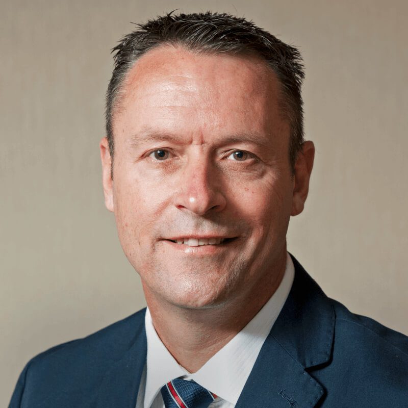 A middle-aged man with short dark hair is wearing a blue suit jacket, white shirt, and a striped tie. He is smiling slightly and is posed against a plain, light-colored background.