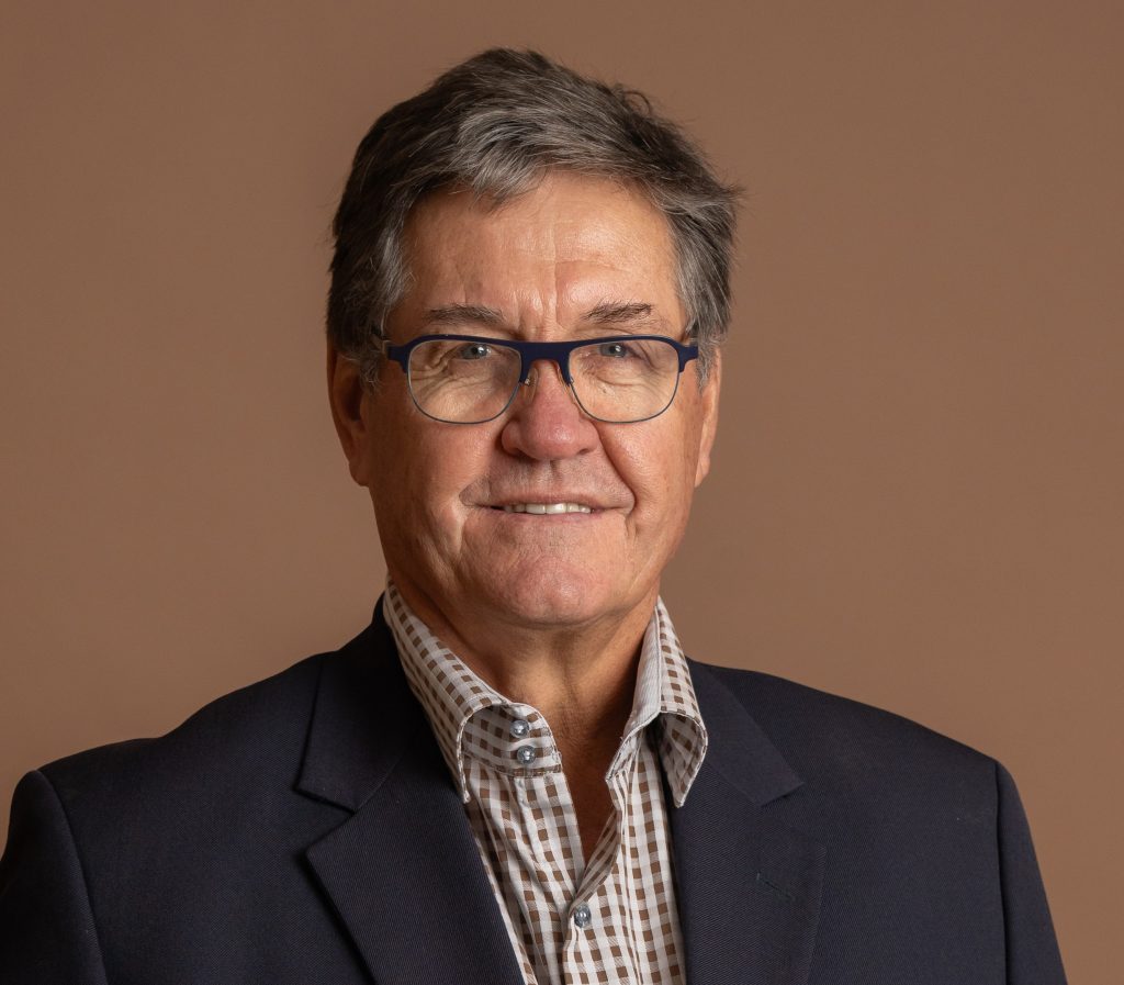 A middle-aged man with short gray hair, wearing glasses, a dark blazer, and a checked shirt, smiles slightly against a plain brown background.