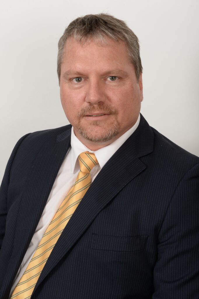 A man with short hair and a beard is wearing a dark pinstripe suit, a white shirt, and a yellow striped tie. He is sitting against a plain white background, looking directly at the camera.