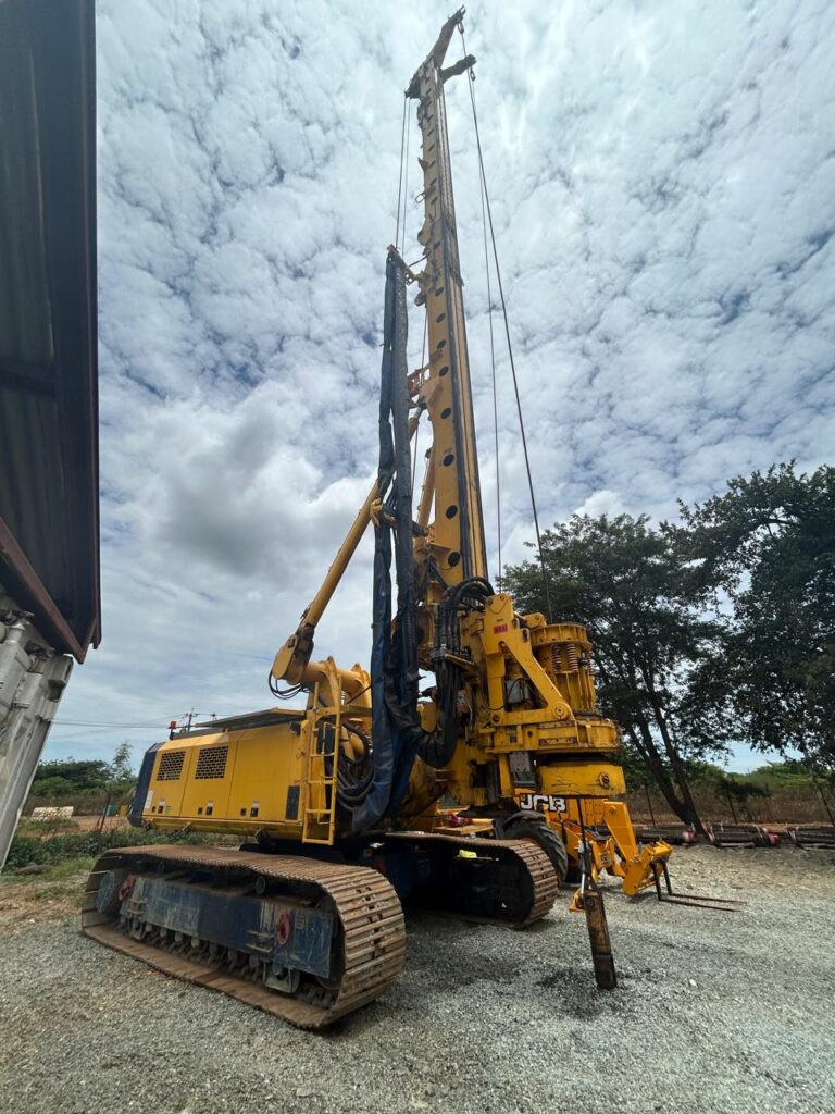 A large yellow drilling rig stands on a gravel surface under a cloudy sky. The heavy machinery has a tall vertical drill, caterpillar tracks, and various hydraulic components. Trees and other equipment can be seen in the background.