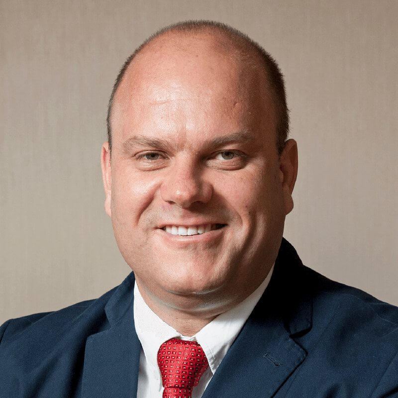 A smiling man with a bald head is wearing a navy blue suit, a white shirt, and a red tie. He is posing in front of a plain, light-colored background.