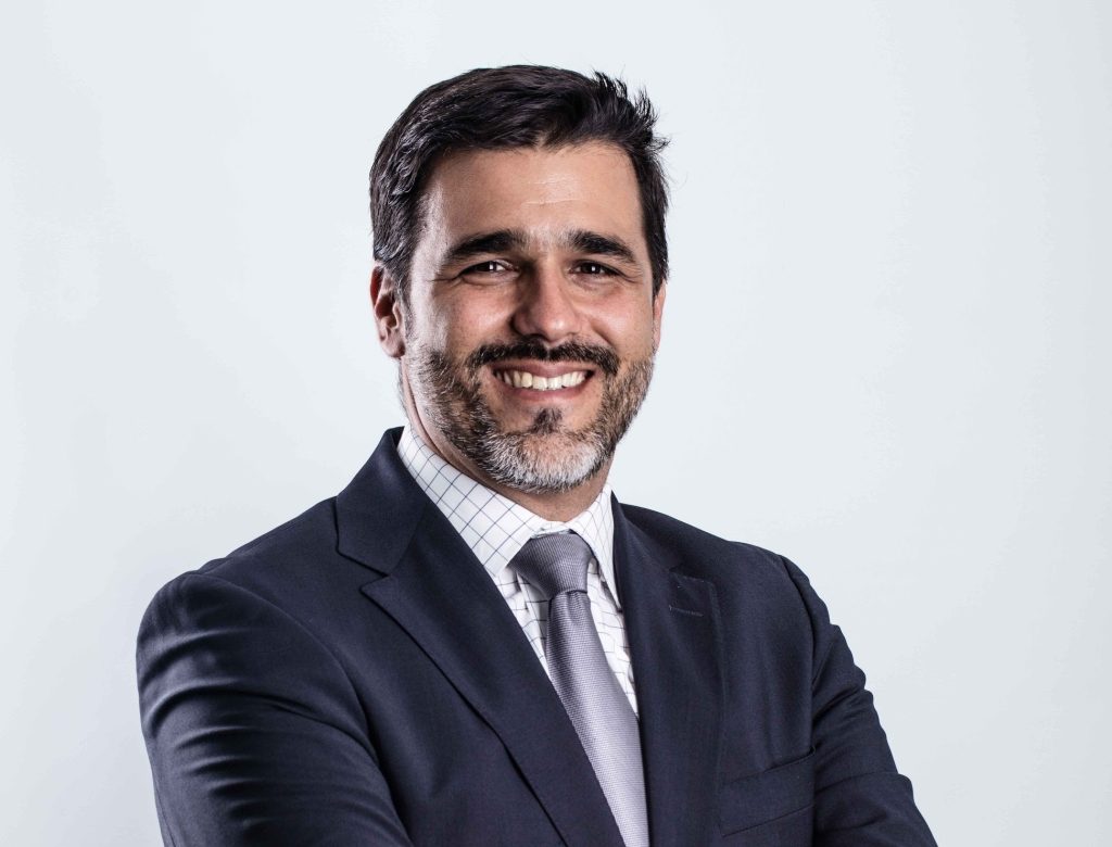 A man in a dark suit, light shirt, and tie stands smiling with arms crossed against a plain light background. He has short dark hair, a beard, and mustache.