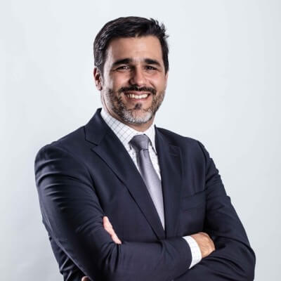 A man with short dark hair and a beard, wearing a dark suit, light-colored tie, and checkered dress shirt smiles at the camera with his arms crossed against a plain light grey background.