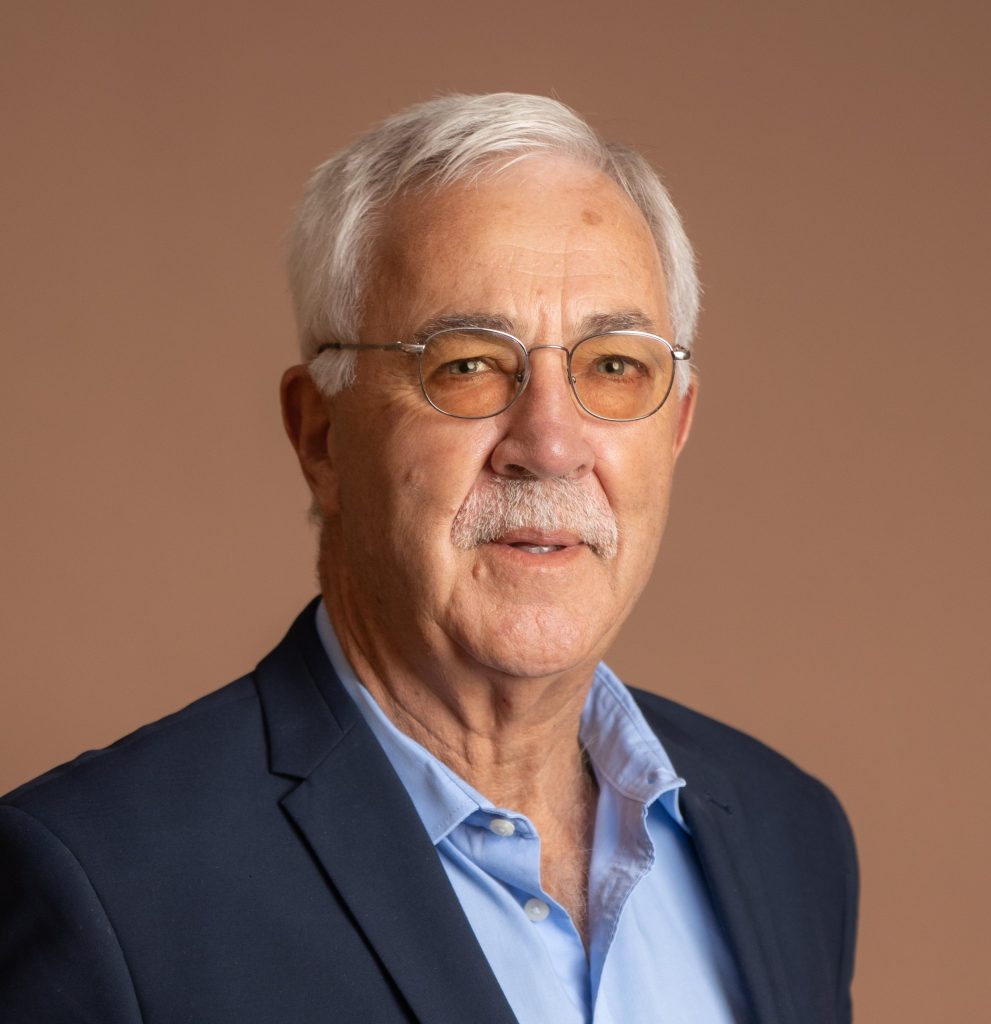 An older man with short gray hair, glasses, and a mustache is wearing a blue dress shirt and a dark blazer, posing against a plain light brown background.