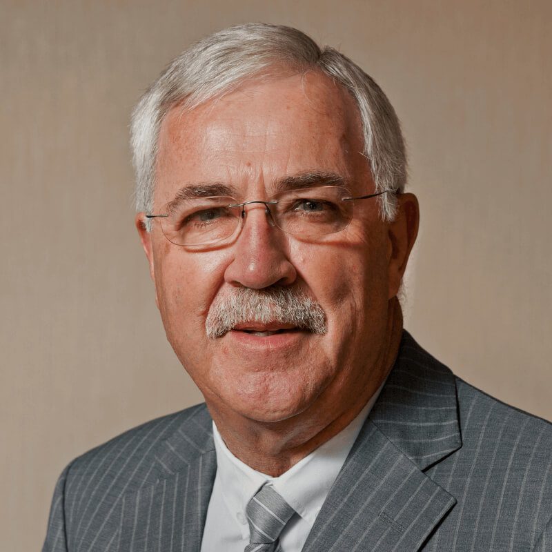 A man with gray hair and a mustache is wearing a gray pinstripe suit, a light gray tie, and glasses. He is standing in front of a neutral-colored background. He has a light complexion and is looking directly at the camera with a slight smile.