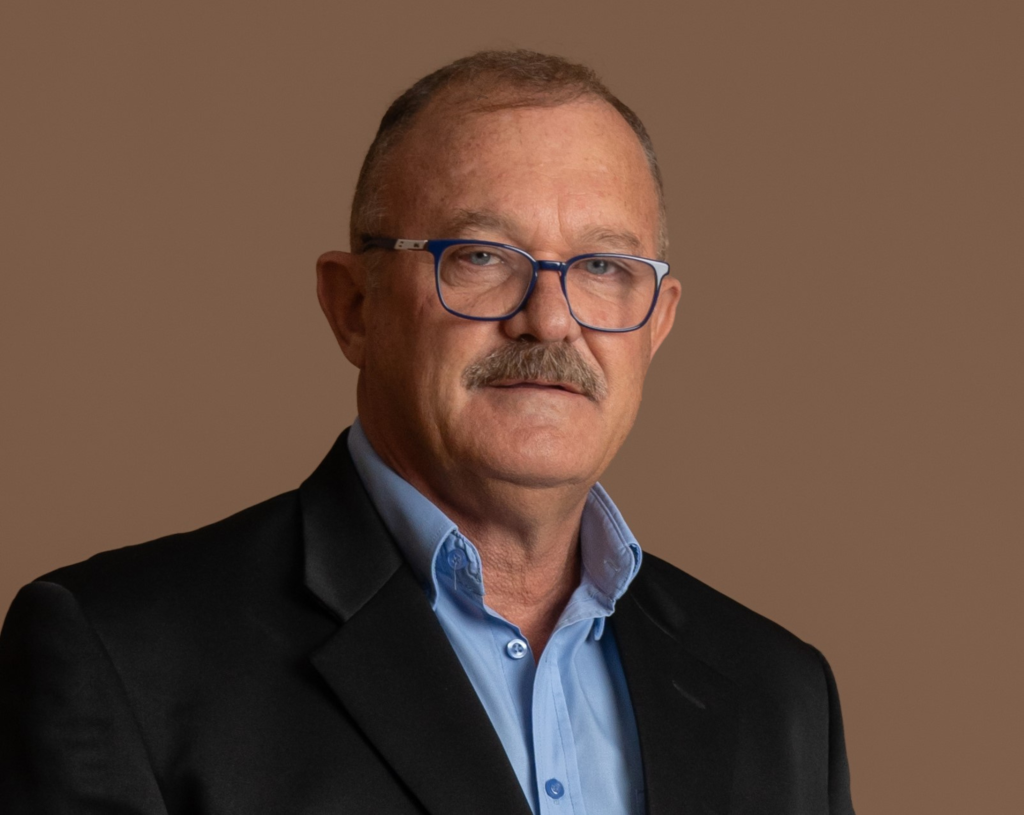 A middle-aged man with short hair, glasses, and a mustache is wearing a black suit jacket over a light blue shirt. He is posing against a plain brown background.