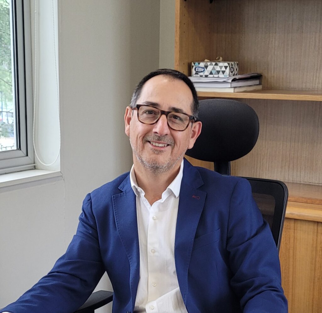 A smiling man with short dark hair and glasses is sitting in an office chair. He is wearing a blue blazer and a white shirt. Behind him are wooden shelves with a few stacked books and a box, and there is a window to his left.