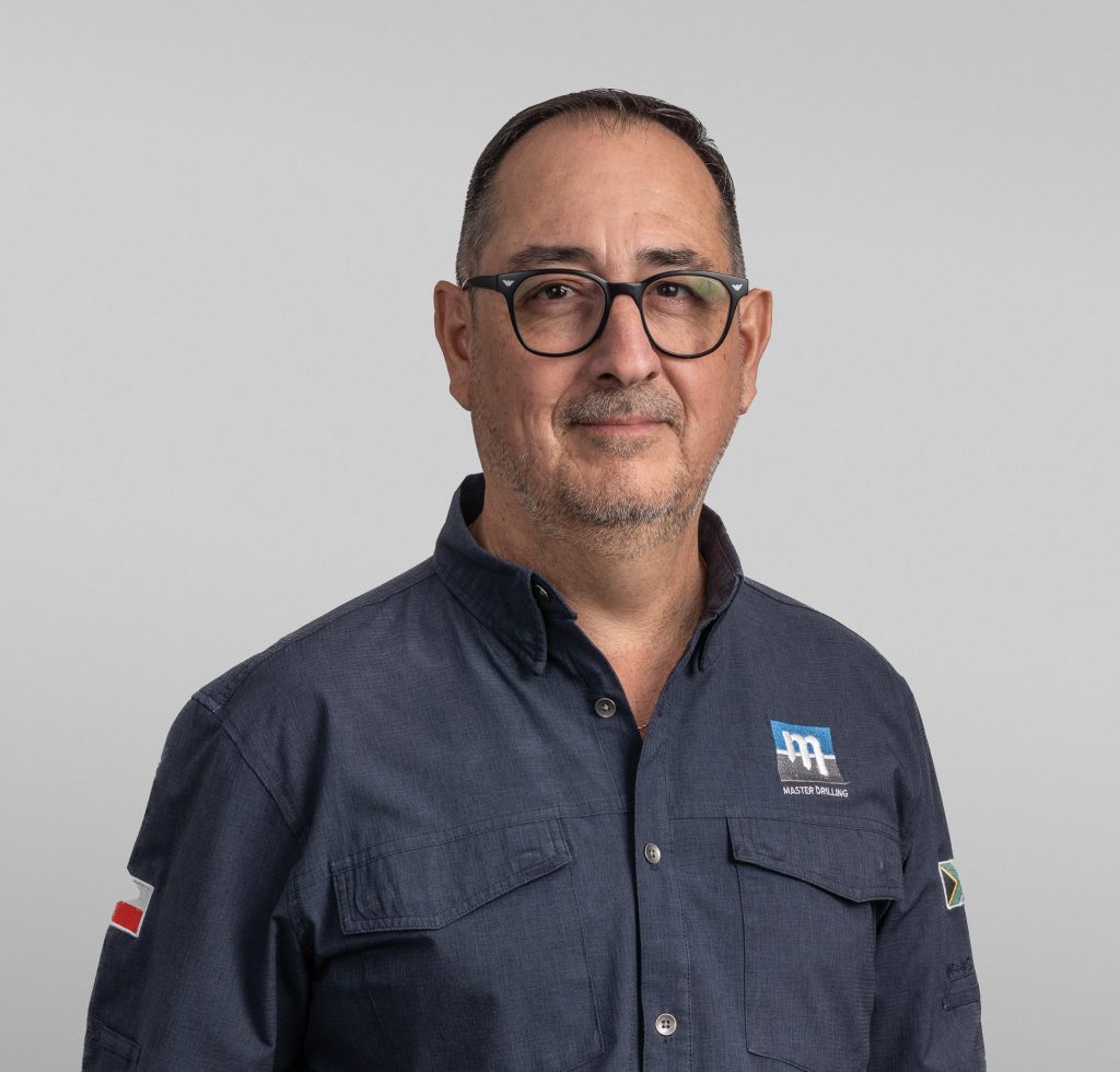 A middle-aged man with short dark hair, glasses, and a trimmed beard wearing a blue button-up shirt with a logo, standing against a plain light grey background.