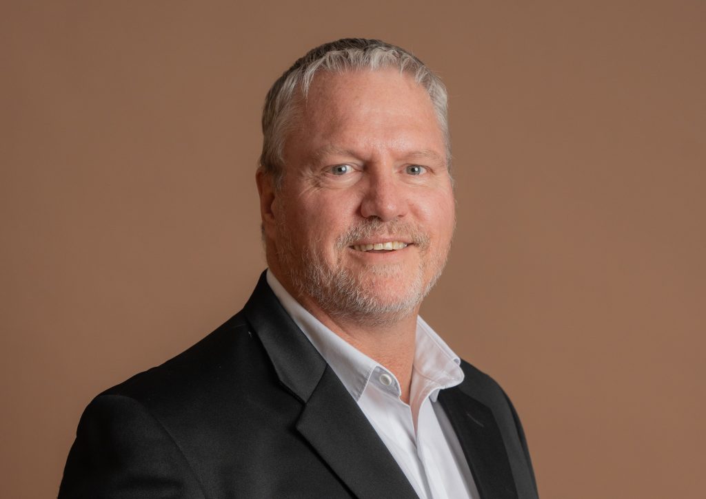 A middle-aged man with short gray hair and a trimmed beard, wearing a black suit jacket and white shirt, smiling in front of a plain brown background.